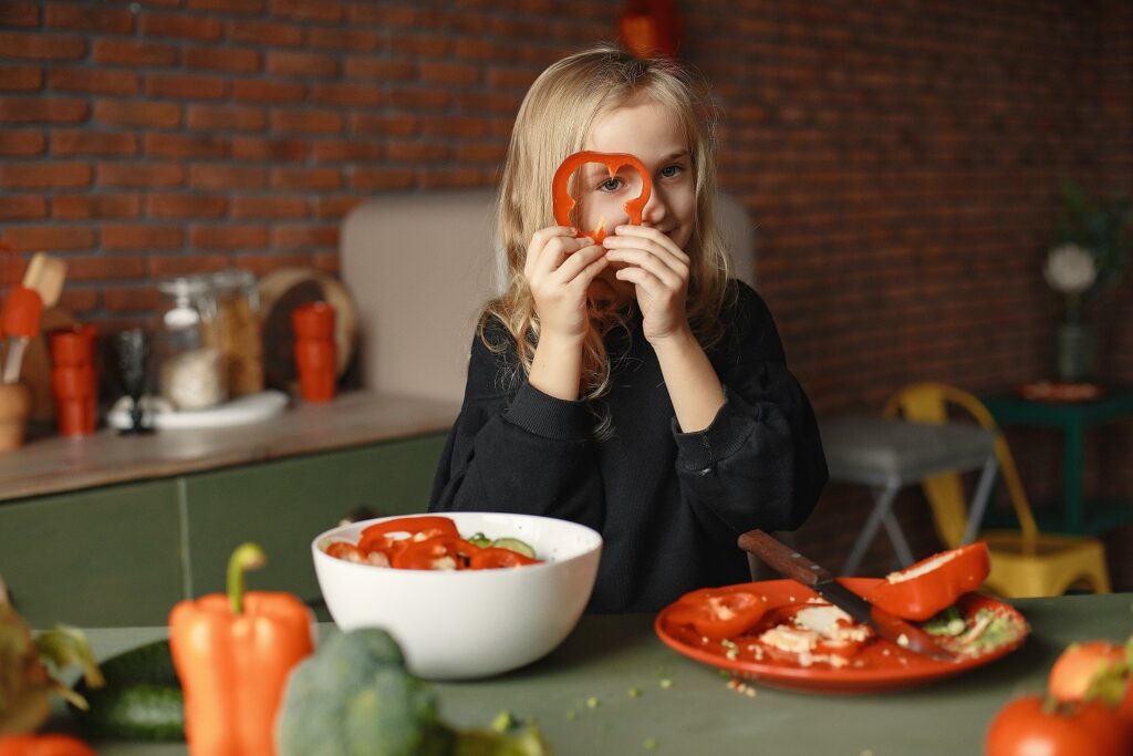 A child holds a red pepper in front of a kitchen table.