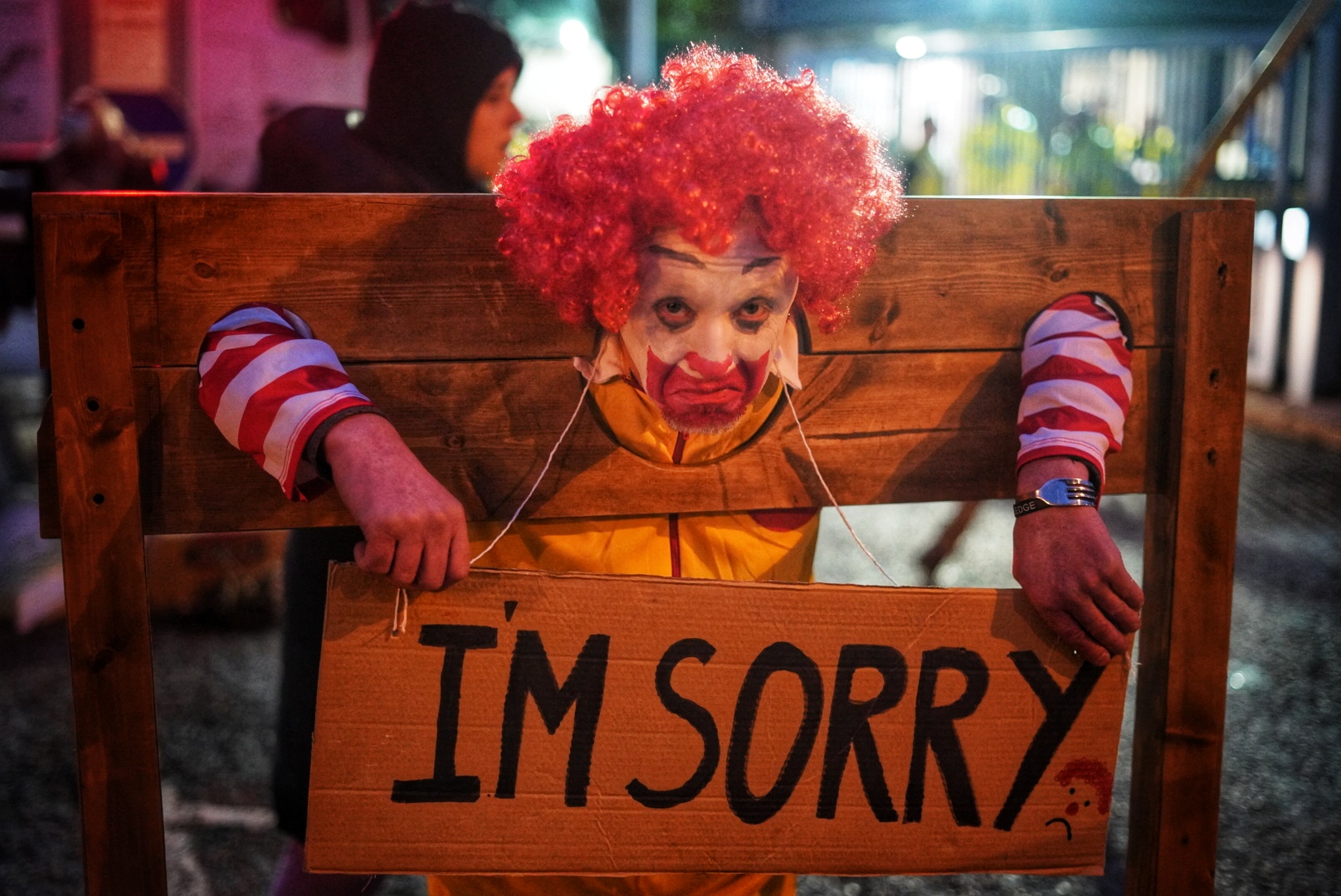 Photo of Alan dressed as Ronald McDonald stuck in a Medieval stock holding a sign that says "I'm sorry"