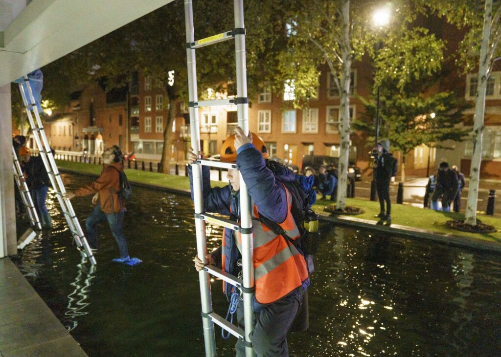 Volunteers with safety equipment wading through water to climb ladders at night to scale the DEFRA building
