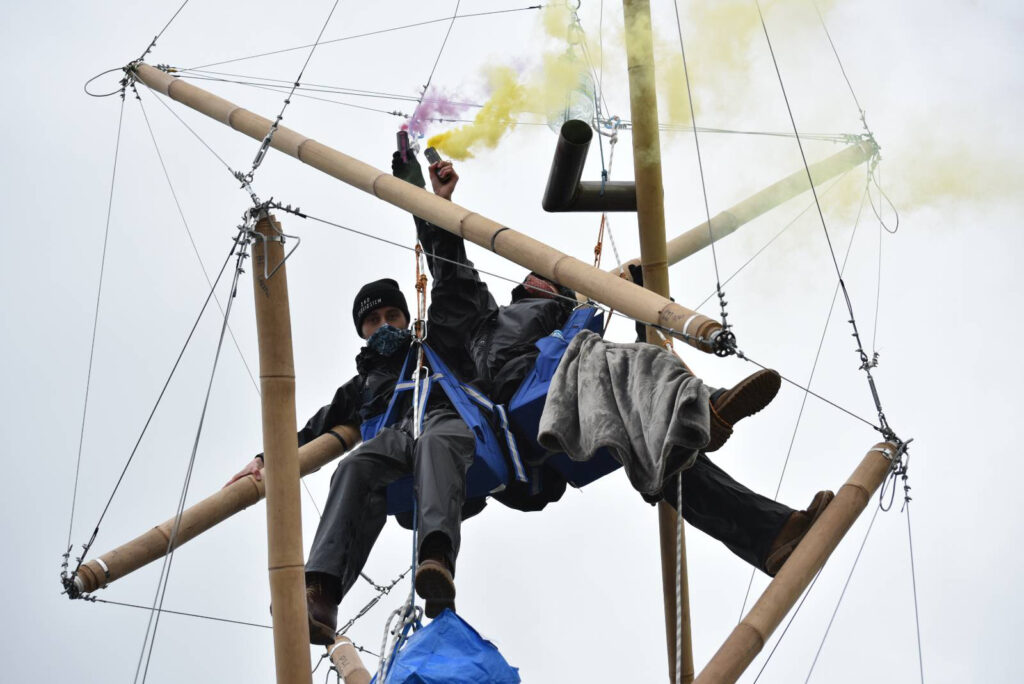 Photo from below of Dillon and another arrestee locked with a bamboo structure high up whilst releasing pink and yellow-coloured smoke