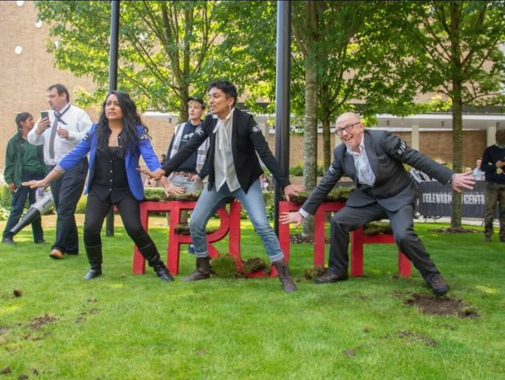 Photo of three people including Alan on grass in tense stances in front of a red structure which spells "truth"