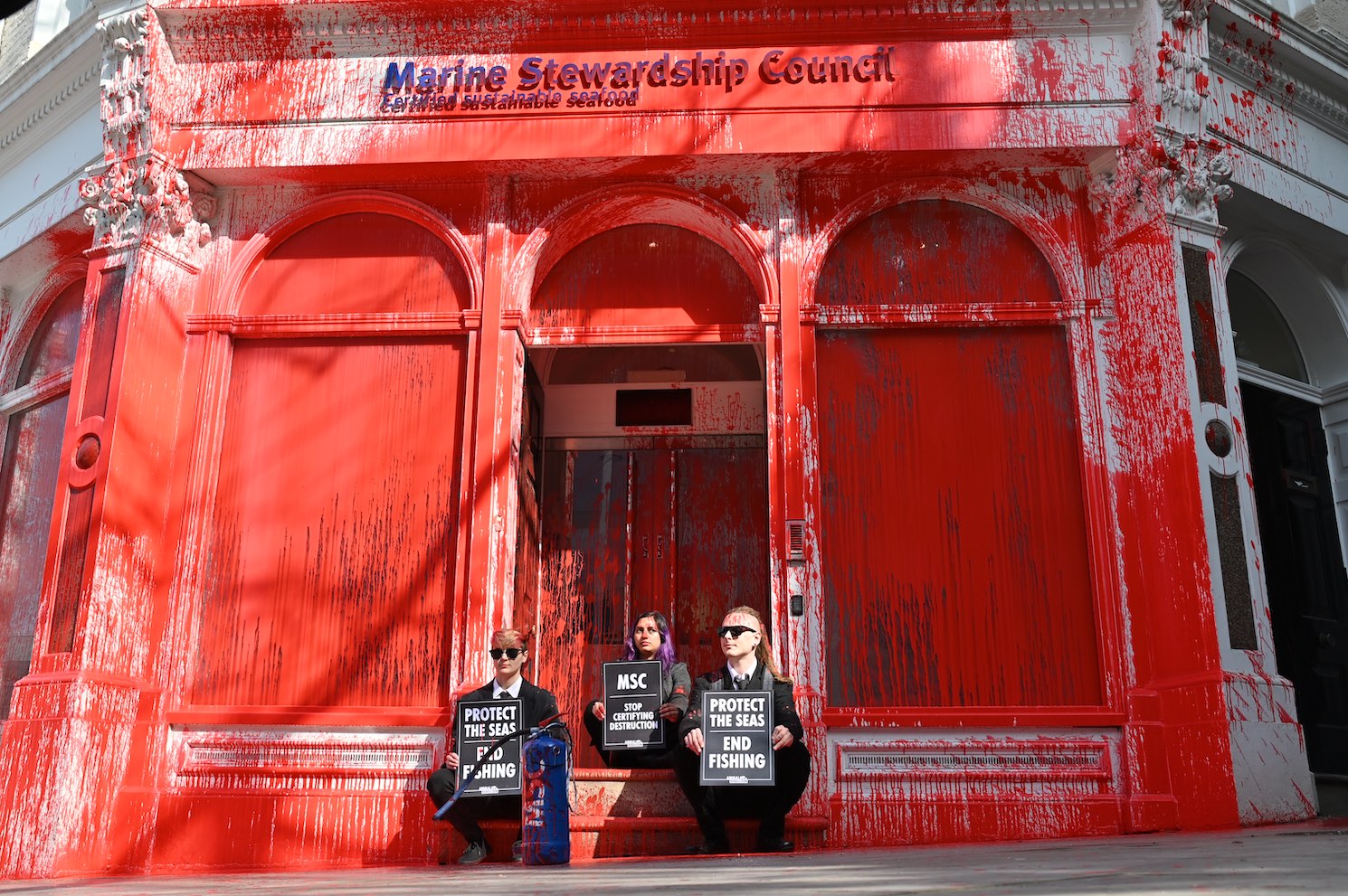 Photo of animal Rebellion activists sit on a step with placards at the entrance to the Marine Stewardship Council. The building is normally white but has been stained bright red.