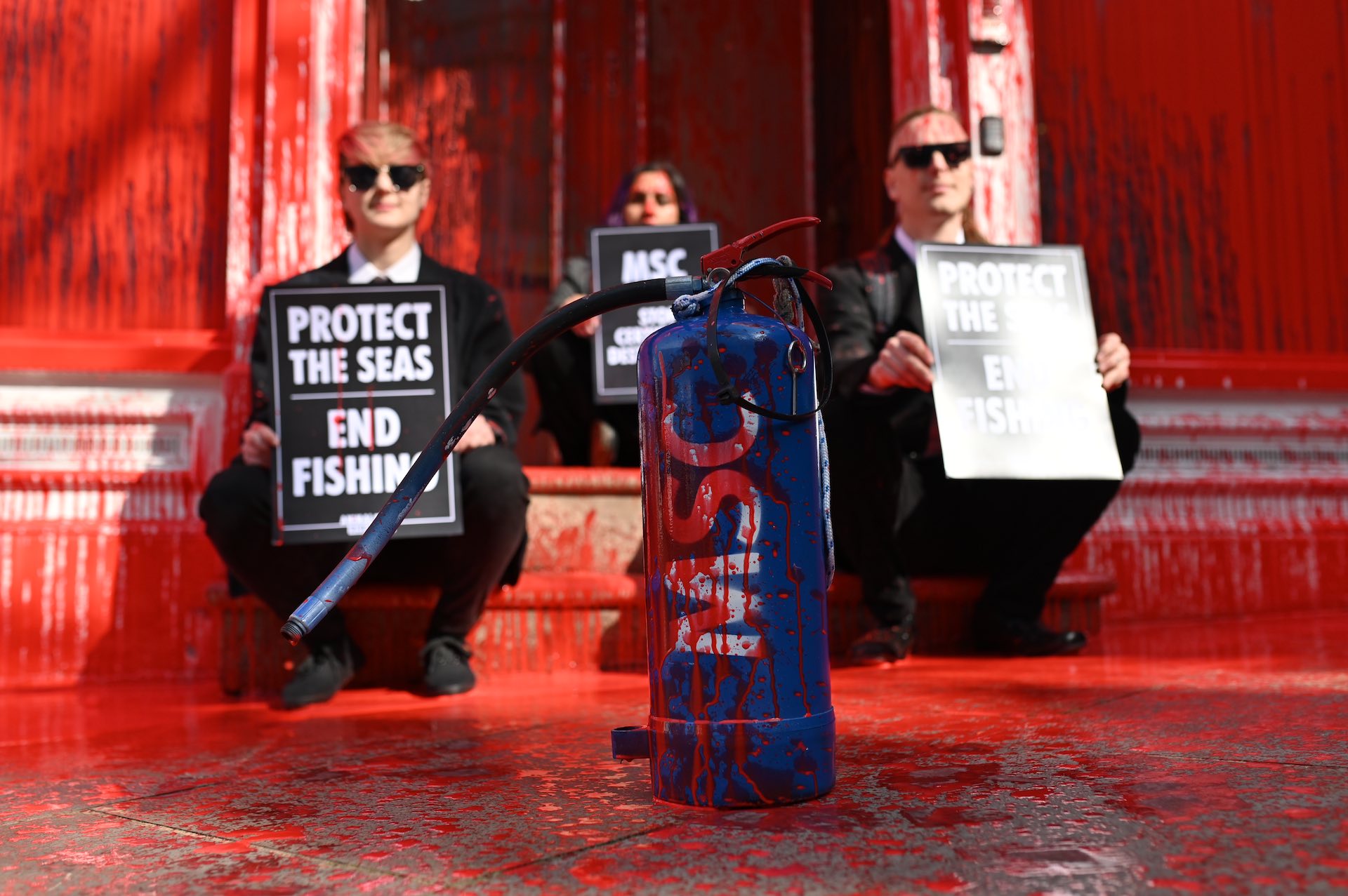 Photo of animal Rebellion activists sitting on a step with placards at the entrance to the Marine Stewardship Council. The building is normally white but has been stained bright red. A blue fire extinguisher with MSC written on it also has been splattered with red paint.